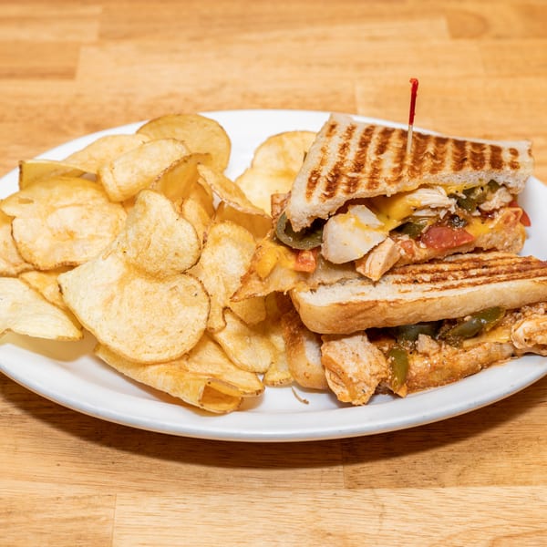 Grilled chicken sandwich with melted cheese, peppers, and tomatoes, served with potato chips on a white plate.
