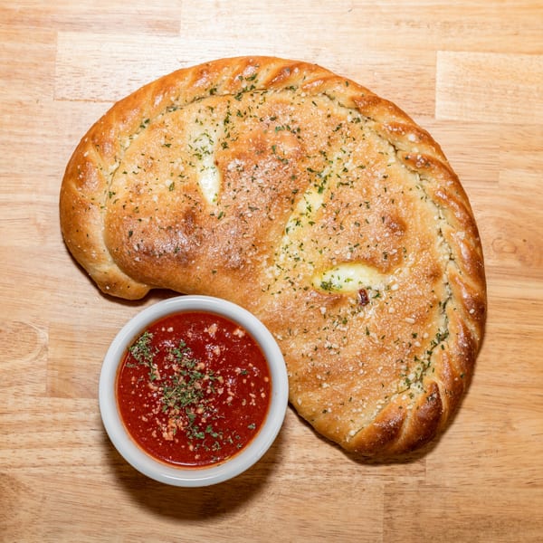A golden-brown calzone sprinkled with herbs and parmesan, next to a bowl of red marinara sauce.