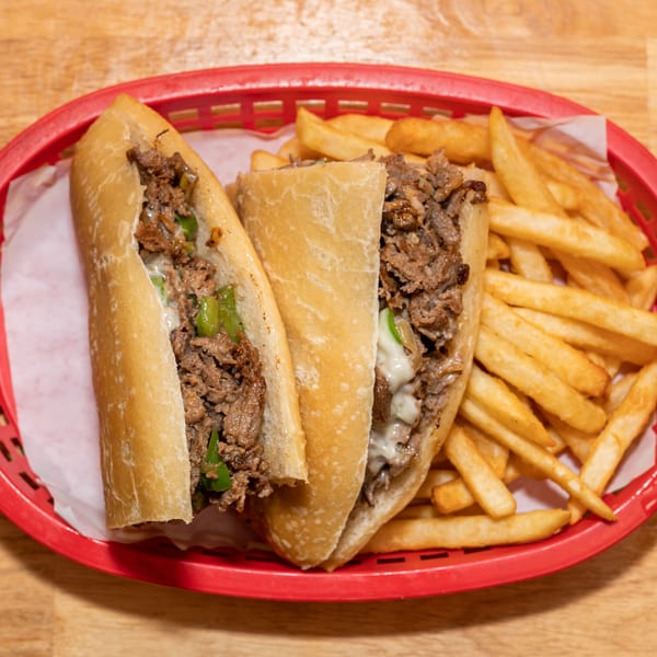 Two cheesesteak sandwiches with green peppers and fries in a red basket on a wooden table.