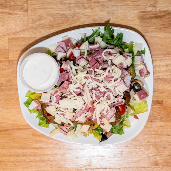 Overhead view of a large antipasto salad with mixed greens, meats, cheeses, and a side of dressing.