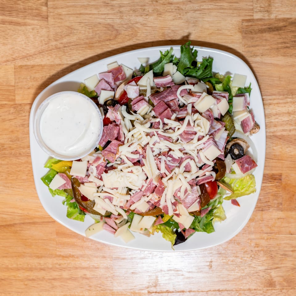 Overhead view of a large antipasto salad with mixed greens, meats, cheeses, and a side of dressing.