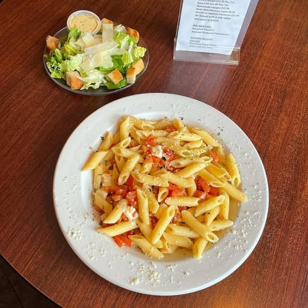 A plate of penne pasta with diced tomatoes and cheese, alongside a Caesar salad and menu.